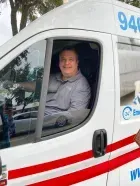 Man smiling in the driver's seat of an ambulance. White vehicle with red stripes; blue emergency number.