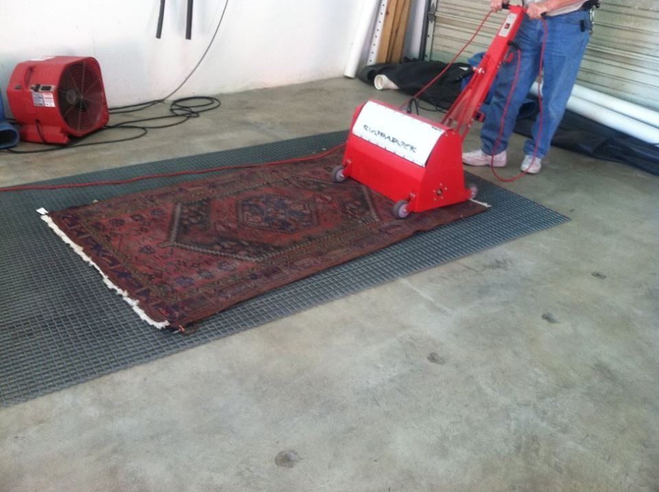 Person using a red rug cleaning machine on a patterned rug in a garage. A fan is visible.