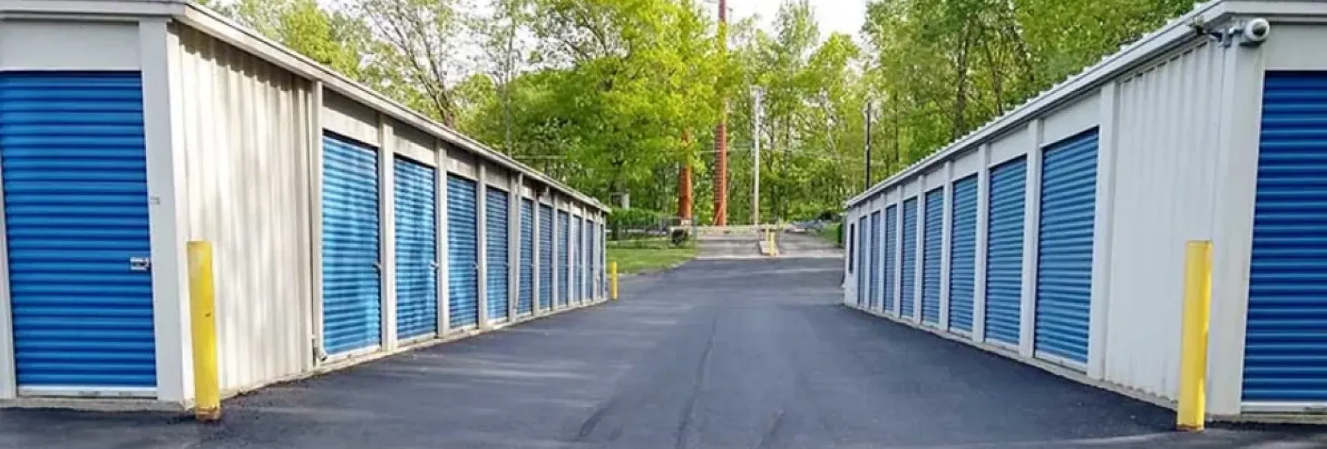 Rows of blue storage unit doors with a paved driveway between them, flanked by yellow safety poles.