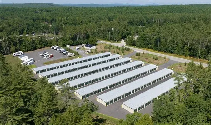 Aerial view of a storage facility with multiple white units. Black asphalt, trees, and houses in the background.