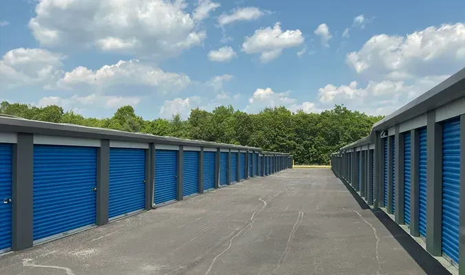 Aerial view of a storage unit facility with rows of individual units, dark asphalt, and a few nearby buildings and a roadway.