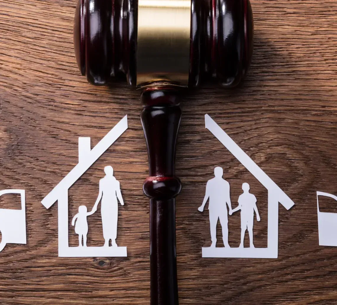 A gavel divides two paper cutouts of split households, each containing figures and a car, on a wood background.