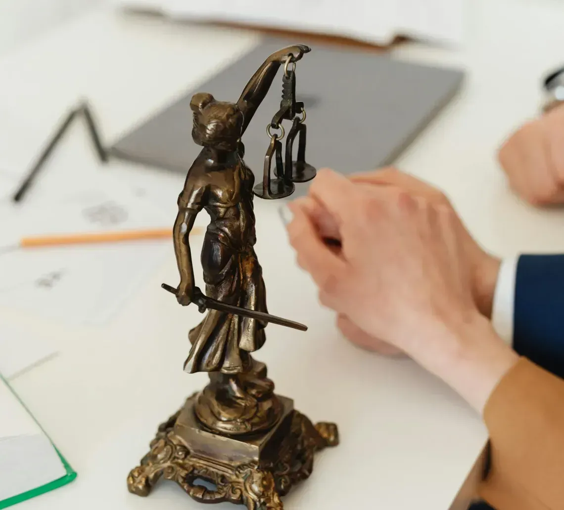 A bronze Lady Justice statue sits on a desk next to a person's hands and a laptop in an office setting.