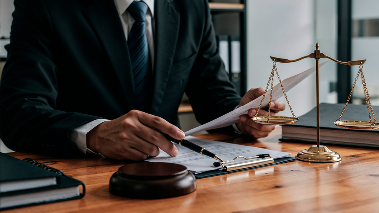 A professional in a suit reviews documents at a desk featuring a wooden gavel and a set of scales of justice.