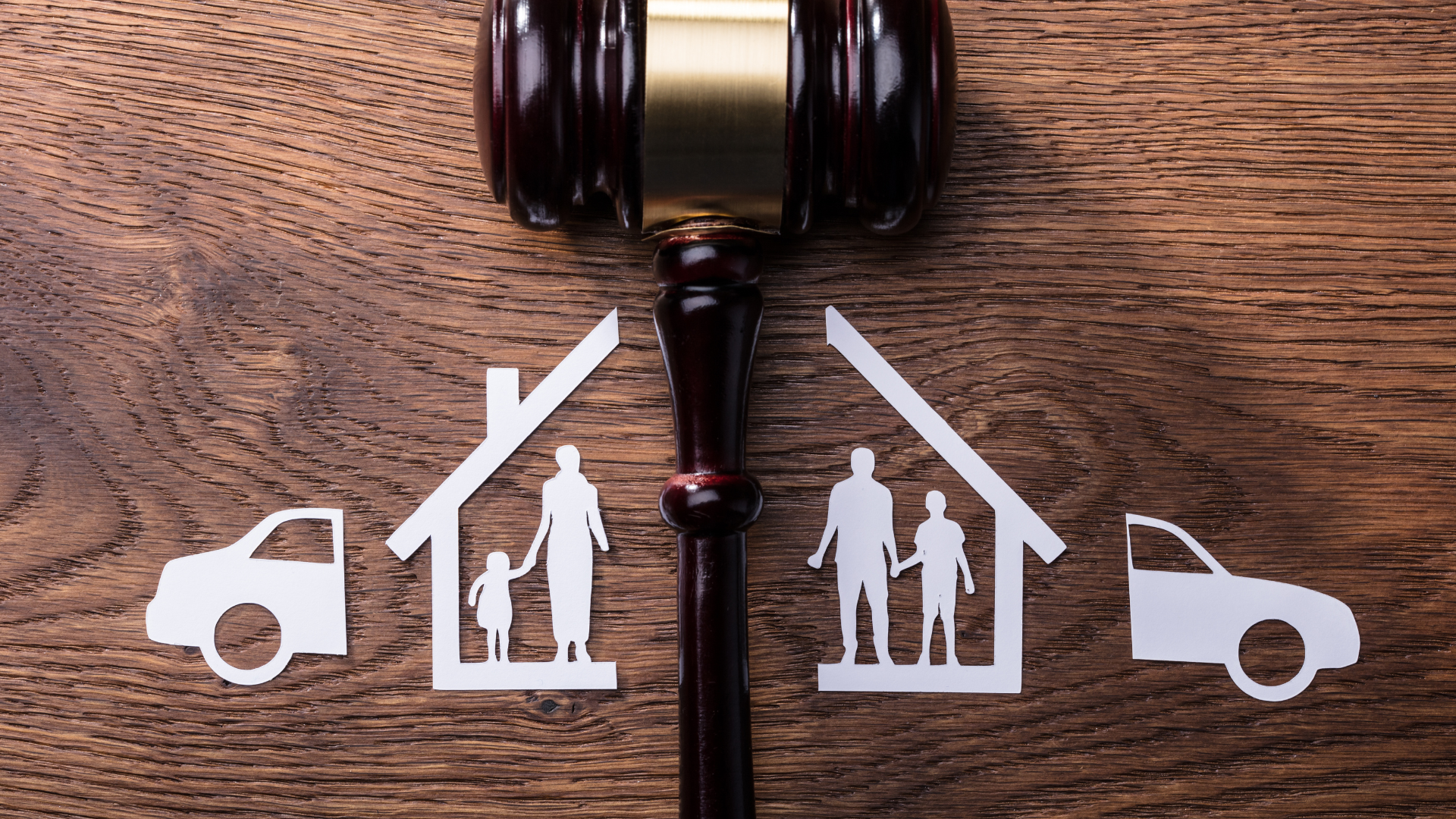 A gavel divides two paper cutouts of split households, each containing figures and a car, on a wood background.