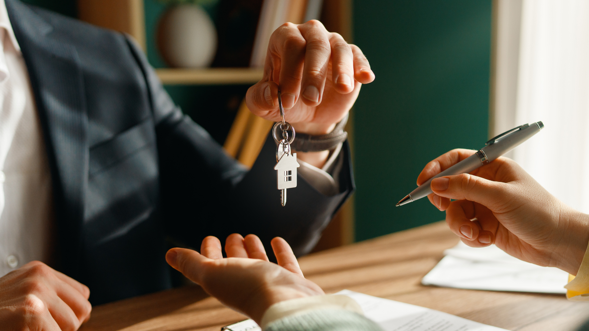 A person in a suit hands house-shaped keys to someone holding a pen over paperwork at a desk.