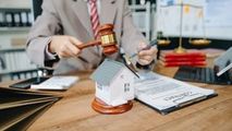 A person in a suit holds a gavel over a miniature house model on a desk with a contract and scales of justice.