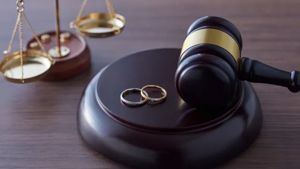 A wooden judge's gavel rests on a sounding block next to two wedding rings, with a small scale of justice in the background.