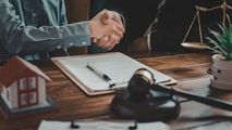 Two people shake hands over a legal document, with a miniature house model, a gavel, and scales of justice on the table.
