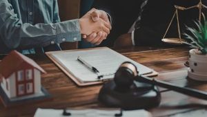 Two people shake hands over a legal document, with a miniature house model, a gavel, and scales of justice on the table.