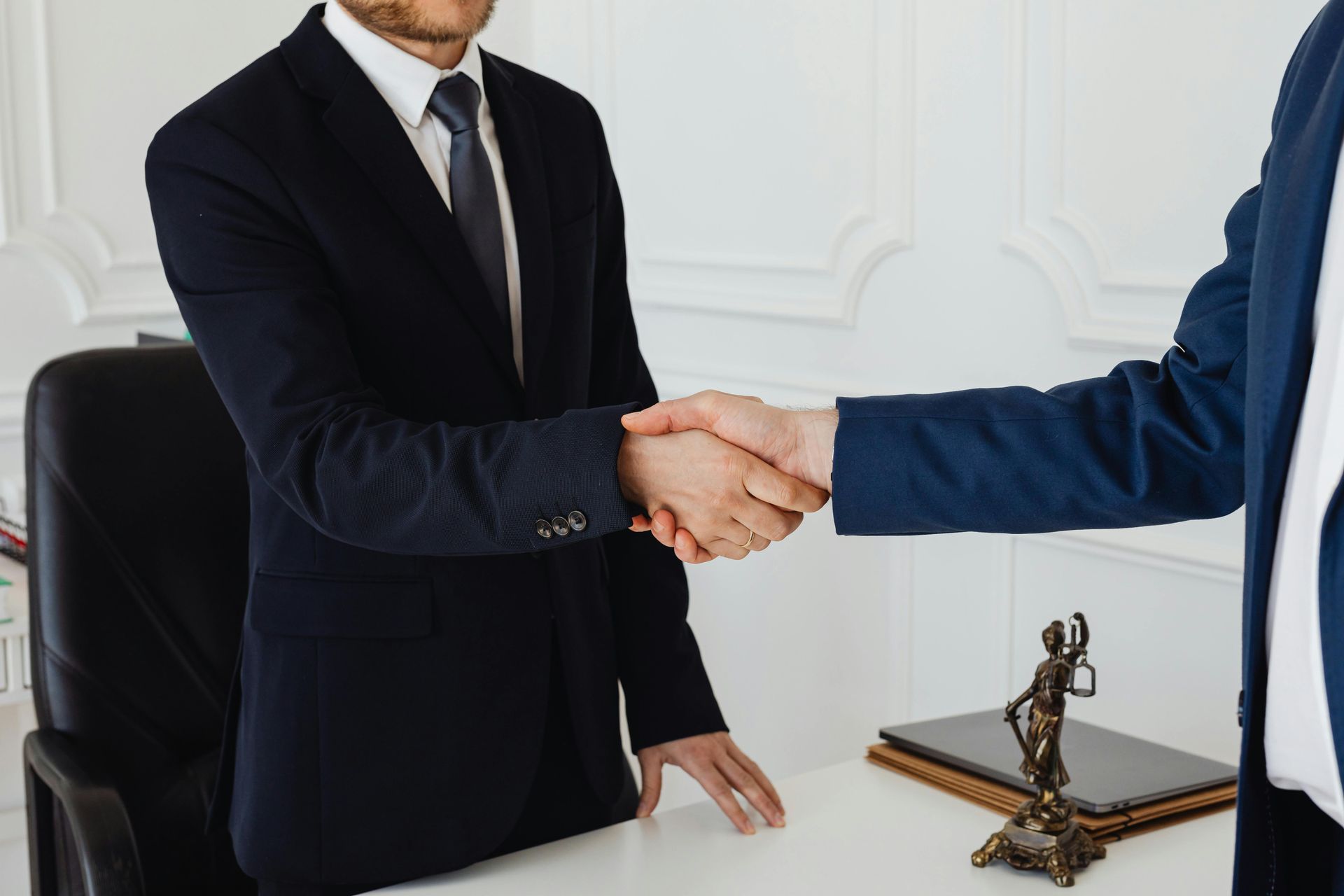 Two professionals in suits shaking hands across a white desk with a small statue of Lady Justice nearby.