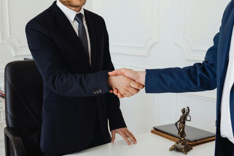 Two professionals in suits shaking hands across a white desk with a small statue of Lady Justice nearby.