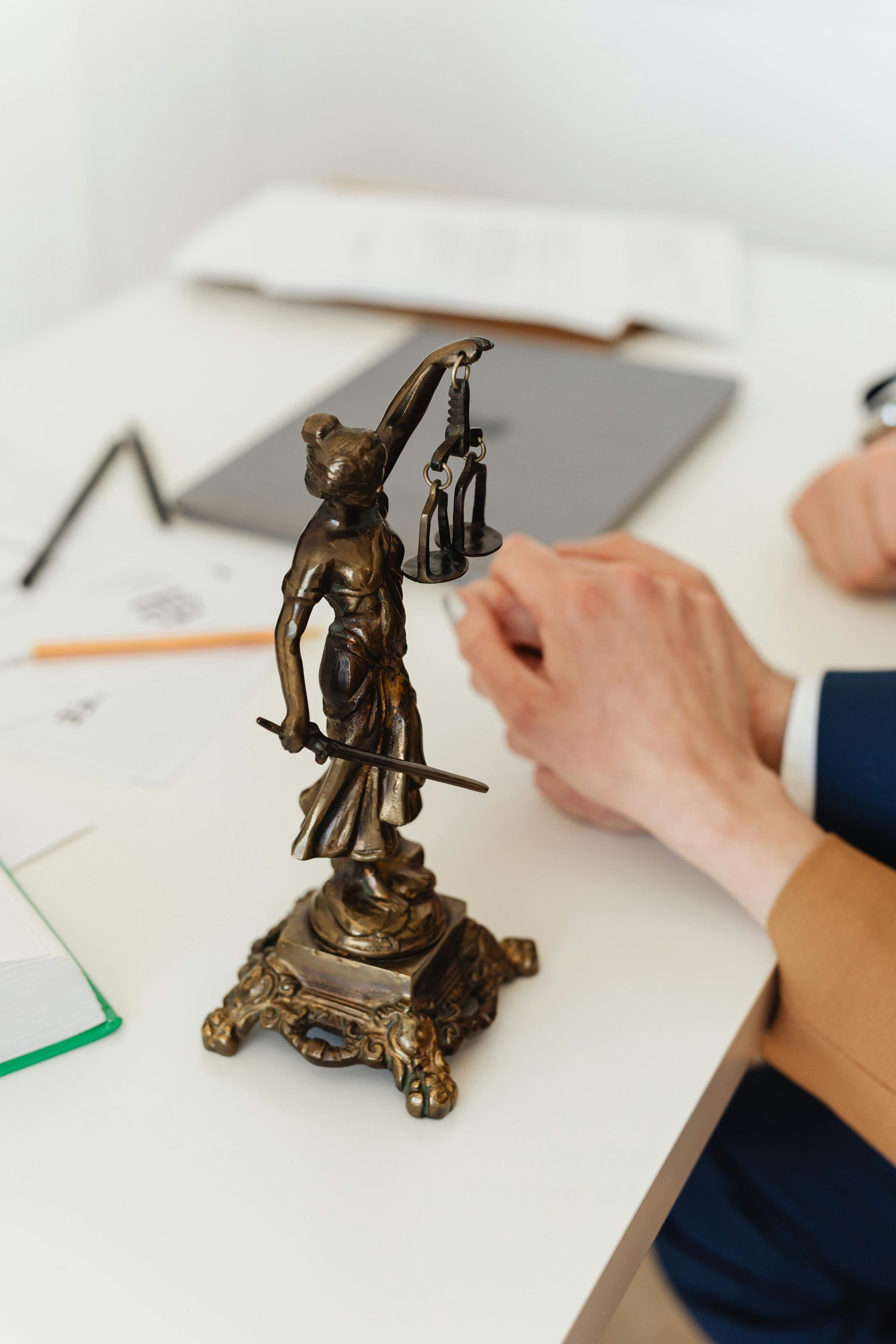 A bronze Lady Justice statue sits on a desk next to a person's hands and a laptop in an office setting.