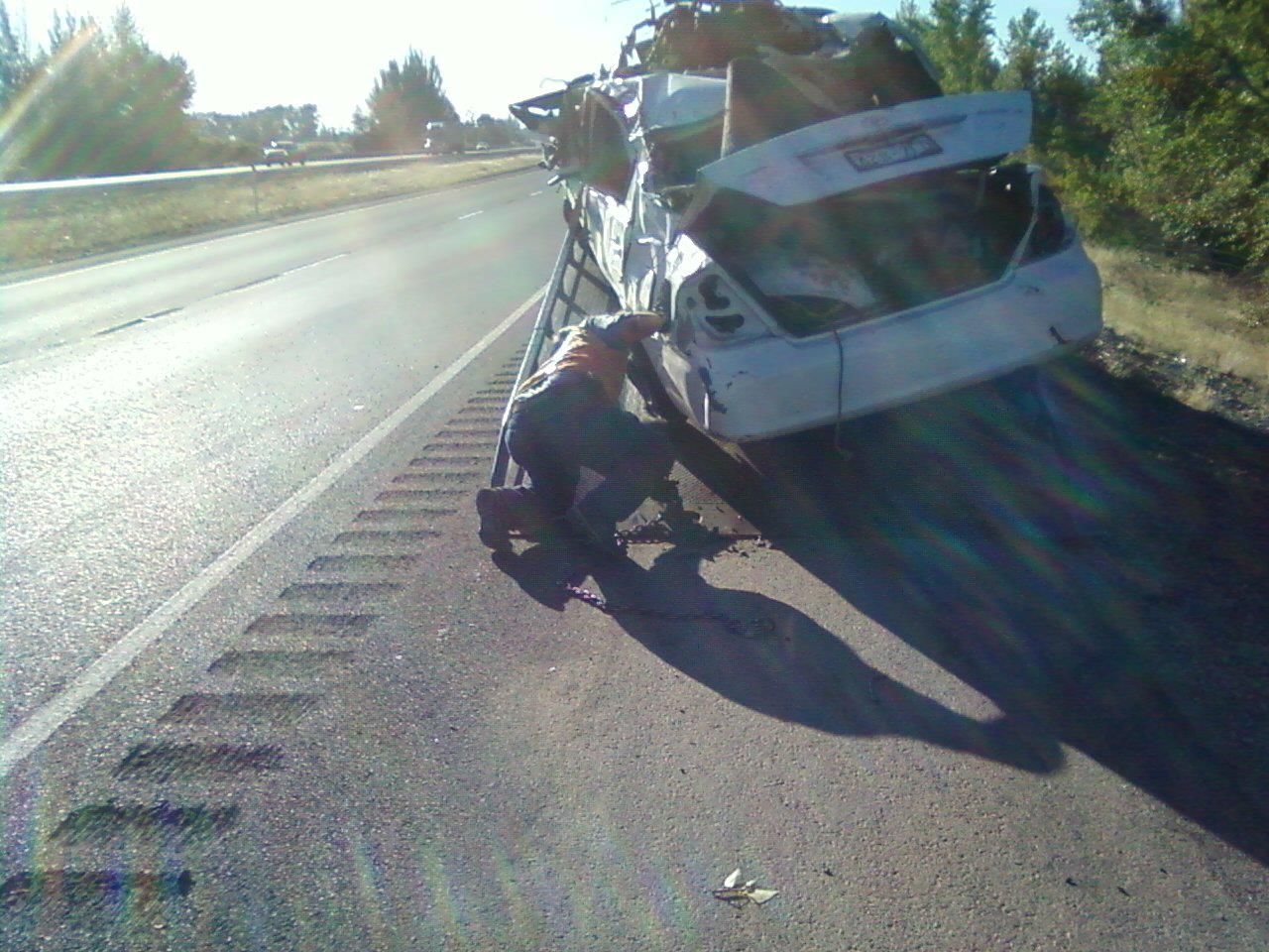 A man is kneeling next to a car that has crashed on the side of the road.