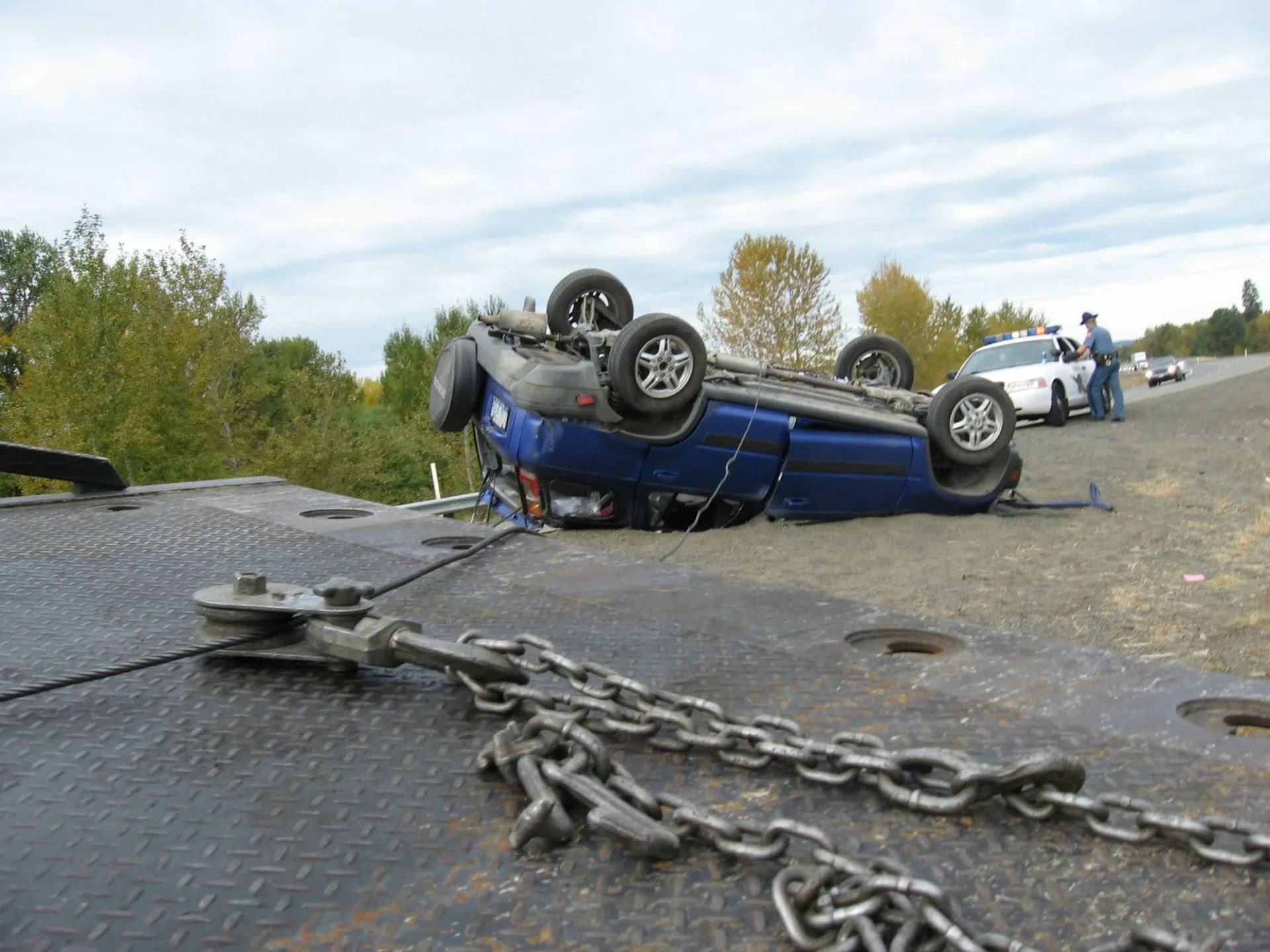 Blue car overturned on tow truck bed, accident scene on roadside.