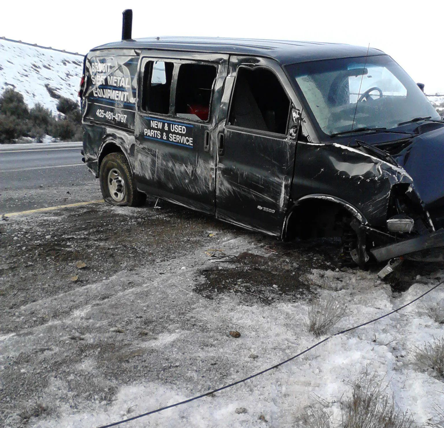 Damaged black van on the side of a road, with snow and a hillside in the background.