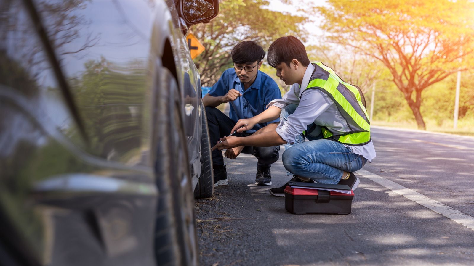 Two people in high-visibility gear examine a flat tire on a vehicle parked on the side of a sunny road.
