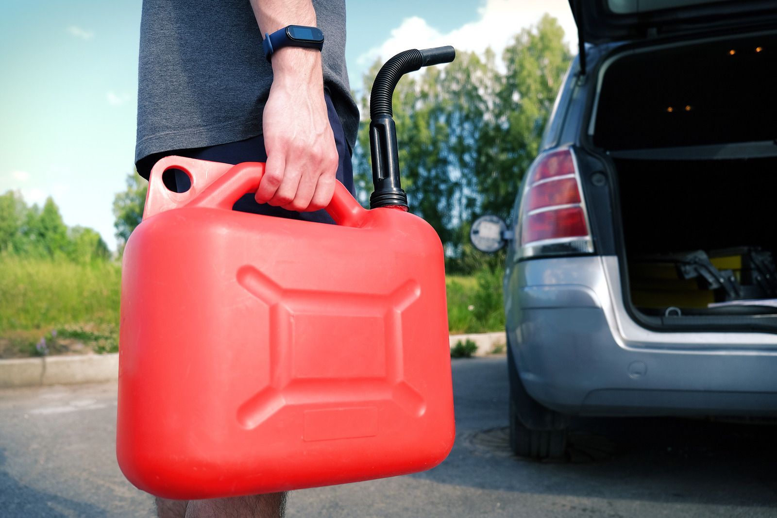 Person holding a red gas can near a car with the trunk open on a sunny day.