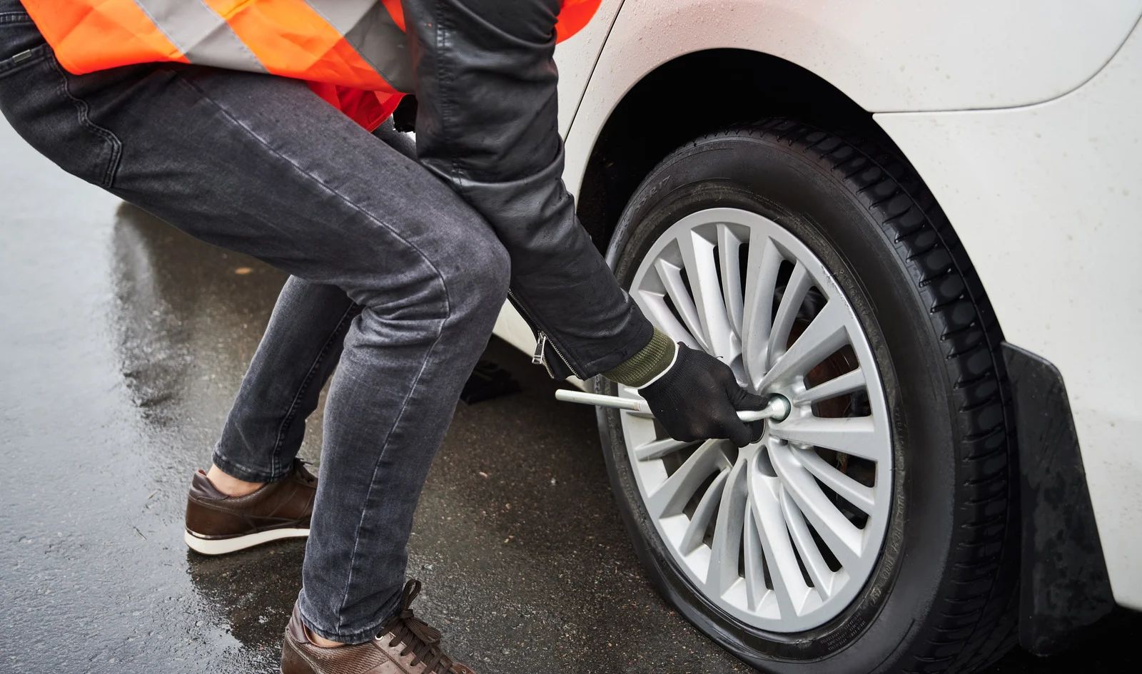 Person changing a tire on a white car, wearing an orange safety vest and black gloves.