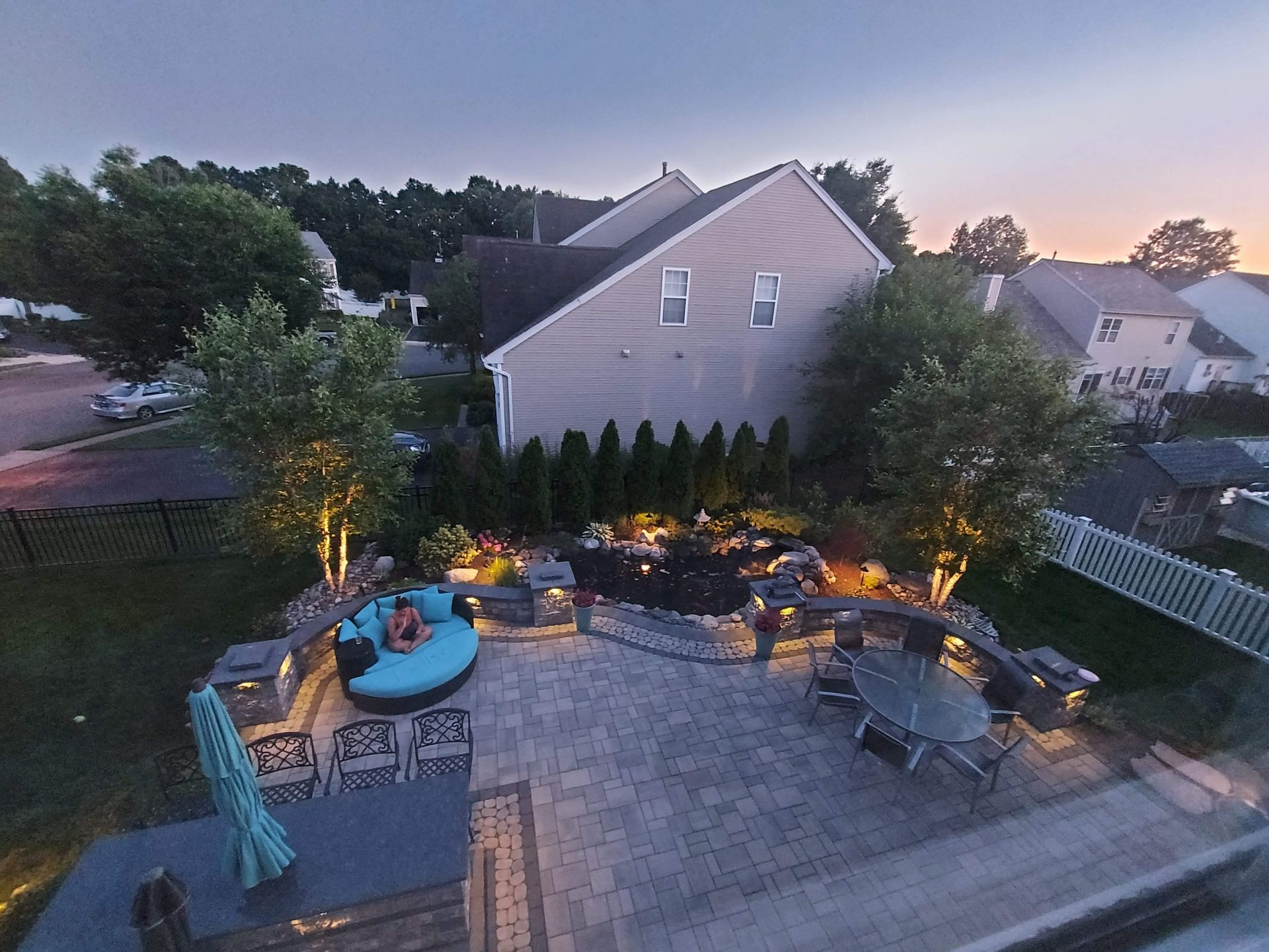 A dusk view of a paver patio featuring a round blue lounge, a dining set, built-in lighting, and a garden landscape.