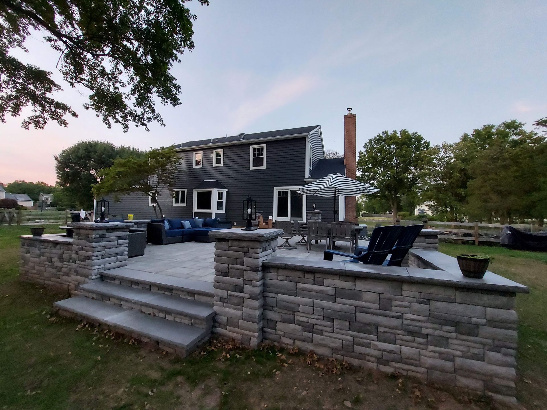 Two people use a compactor on a sand-covered backyard patio area next to a white house. Two people use a compactor on a sand-covered backyard patio area next to a white house.