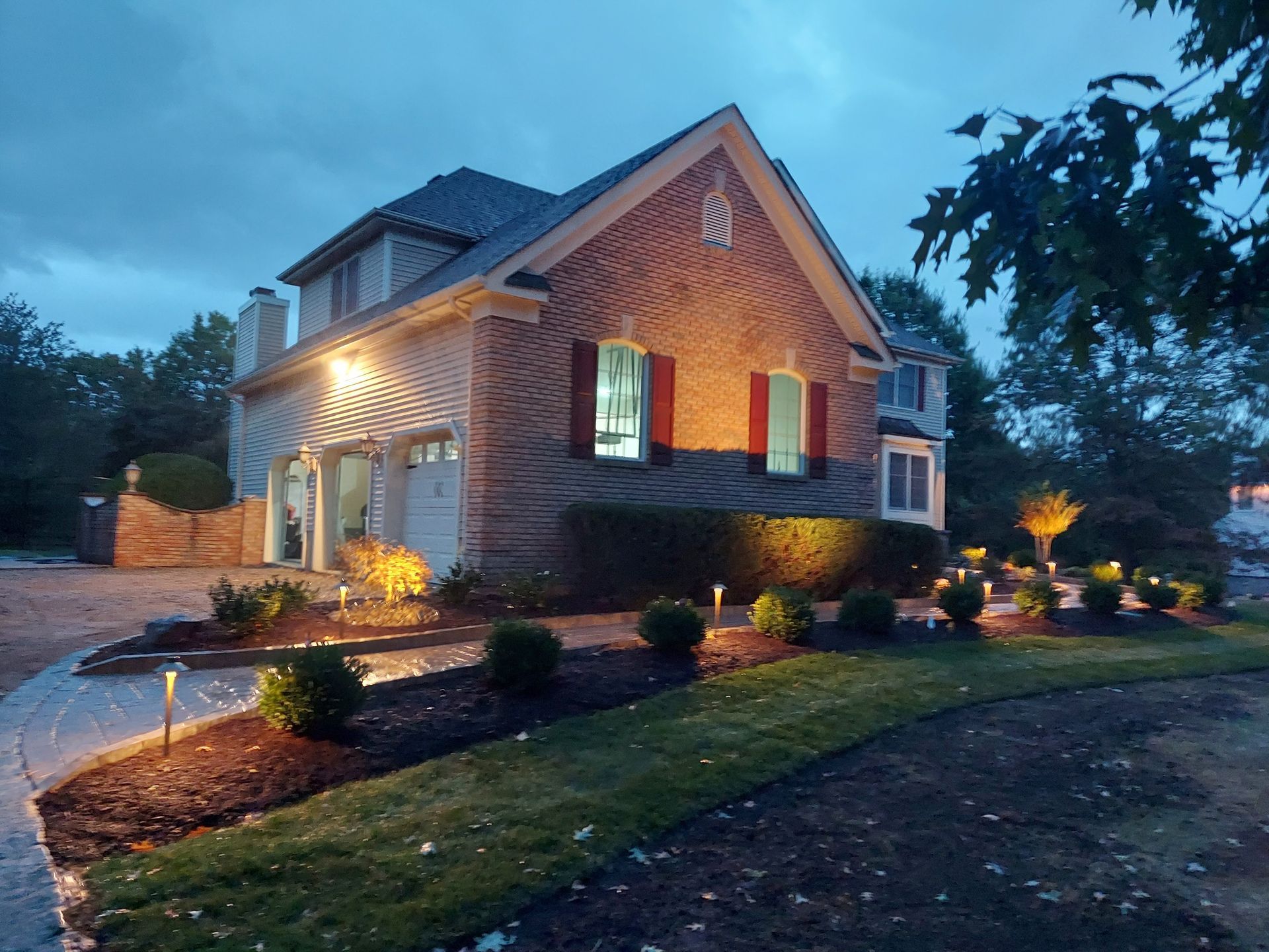 A two-story brick house at dusk, illuminated by glowing warm lights along the driveway and landscaped yard.