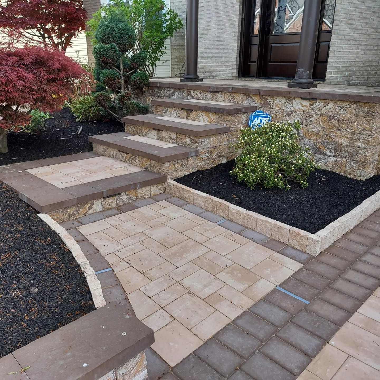 Stone steps with beige pavers and dark borders lead up to a doorway surrounded by red and green landscaping.