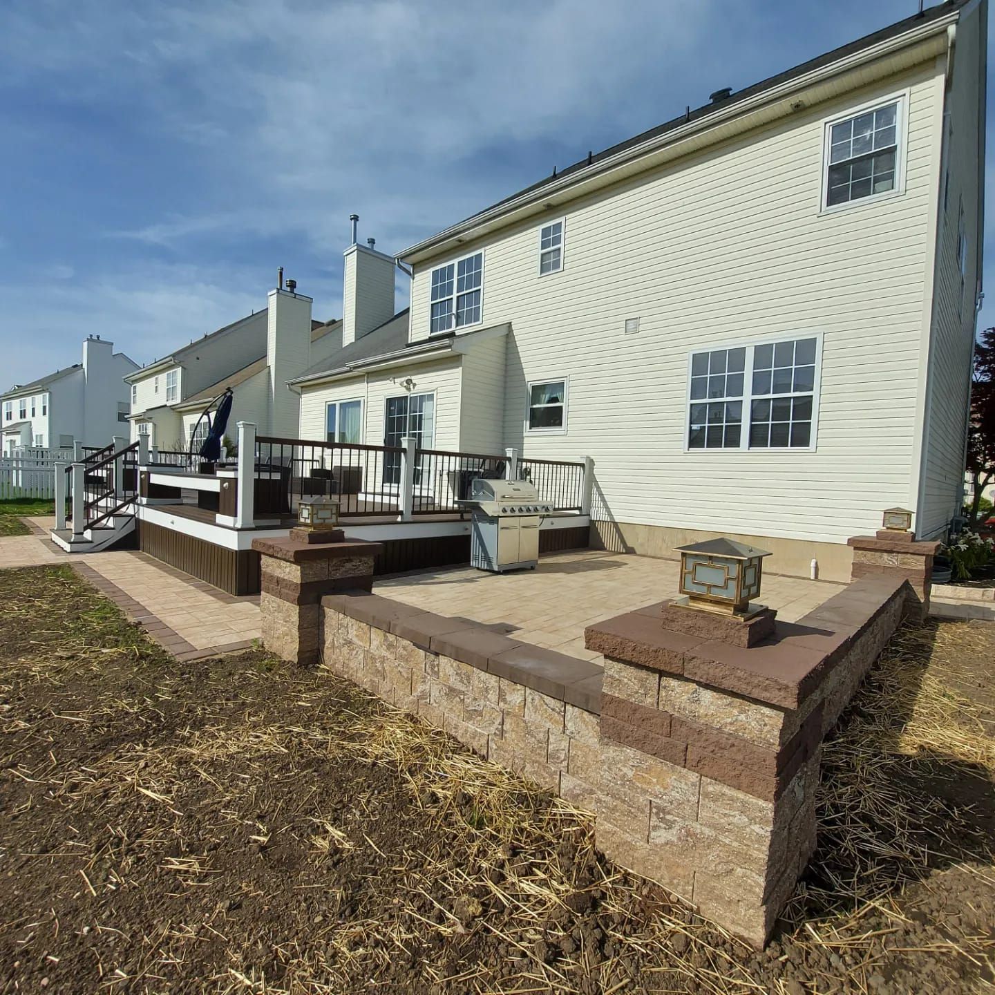 A backyard scene featuring a cream-colored house, an elevated wooden deck, and a stone patio wall with decorative lights.