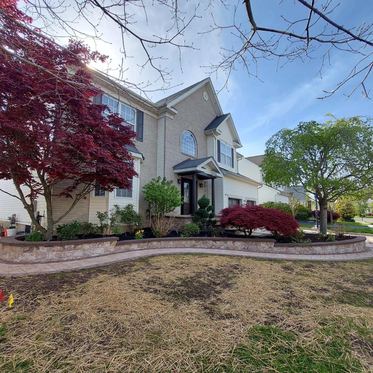 A two-story tan house with a stone retaining wall, front garden beds, a large red maple, and green trees under a blue sky.