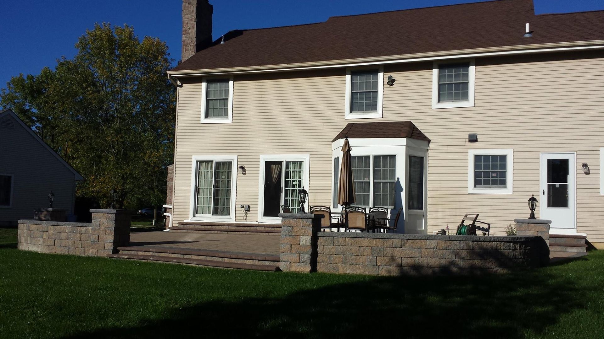 A tan two-story house features a stone patio with retaining walls and a dining set under an umbrella on a sunny lawn.