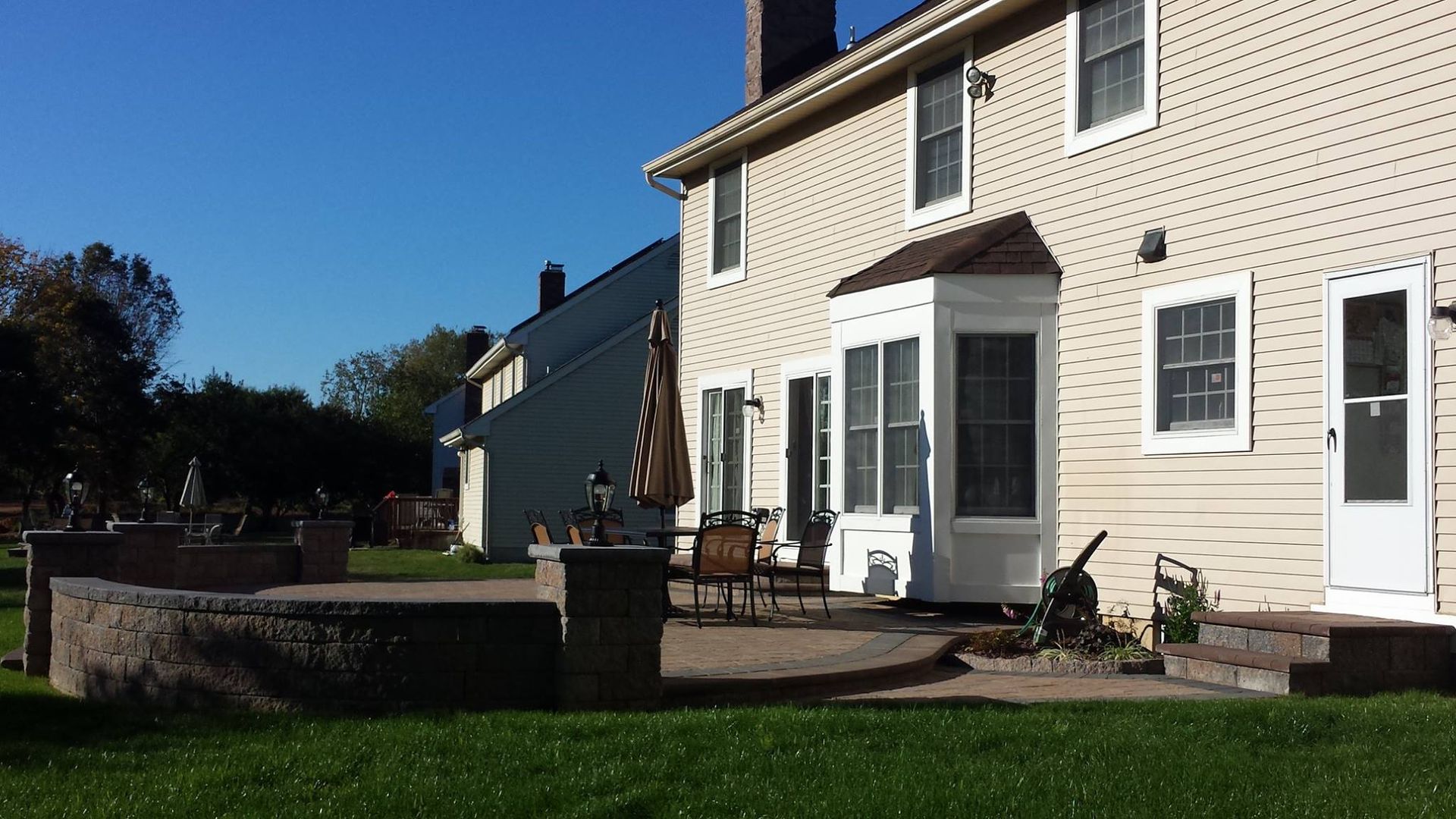 A tan house with a stone patio, low wall, and bay window in a green backyard on a sunny day.
