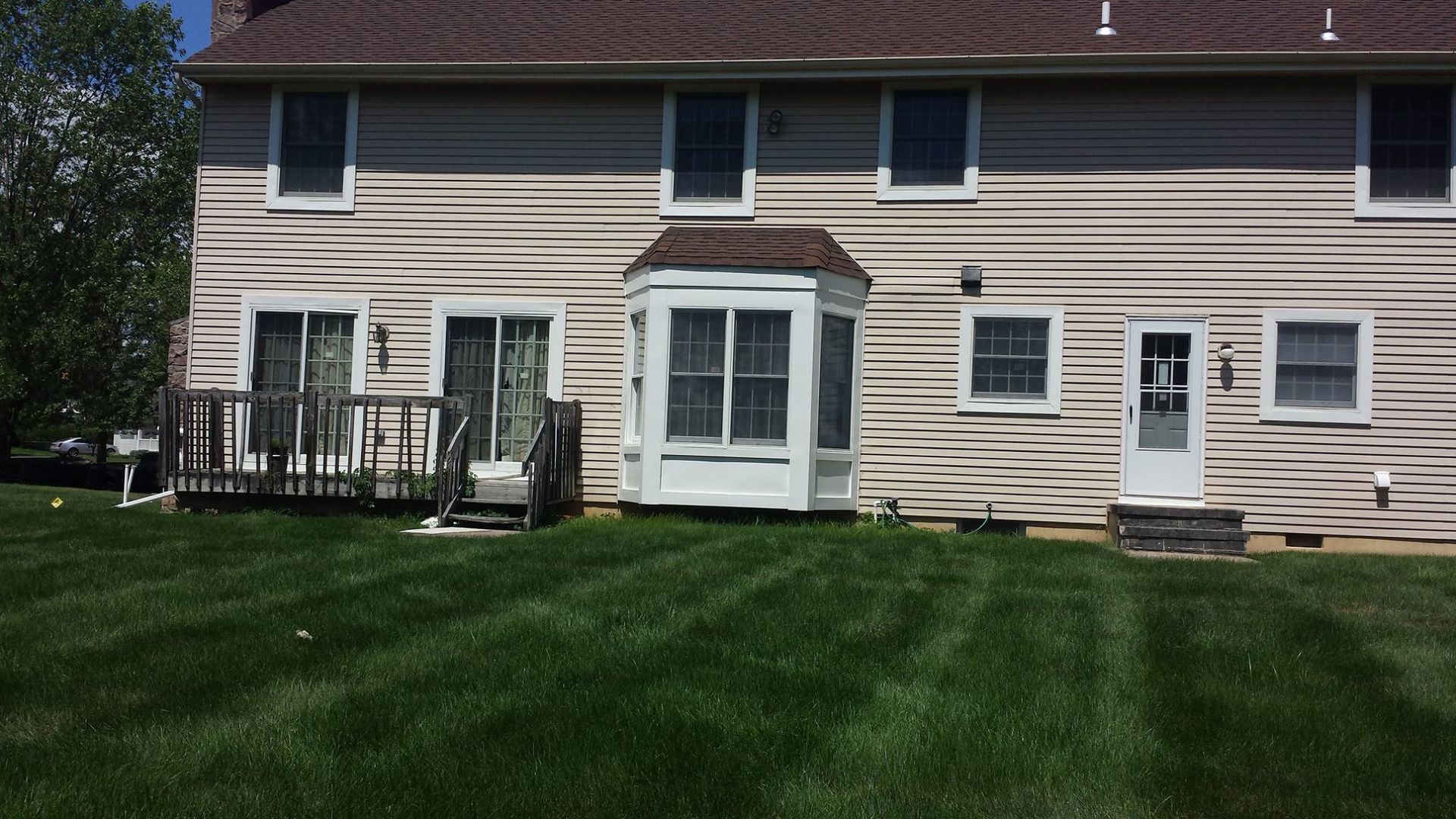 The rear view of a two-story house with beige siding, a wooden deck, a bay window, and a green lawn under a clear sky.