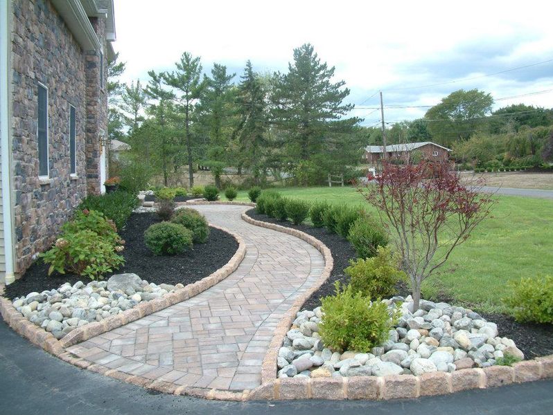 A curved paver walkway bordered by stones and manicured shrubs leads toward a stone-faced house. A curved paver walkway bordered by stones and manicured shrubs leads toward a stone-faced house.