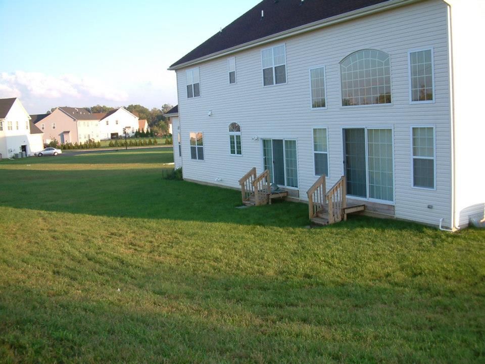 The back of a large white two-story suburban house with wooden steps leading to sliding glass doors, facing a large yard. The back of a large white two-story suburban house with wooden steps leading to sliding glass doors, facing a large yard.