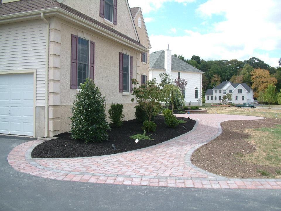 A tan house with a brick paver walkway leading to its front, surrounded by black mulch landscaping and other homes nearby.