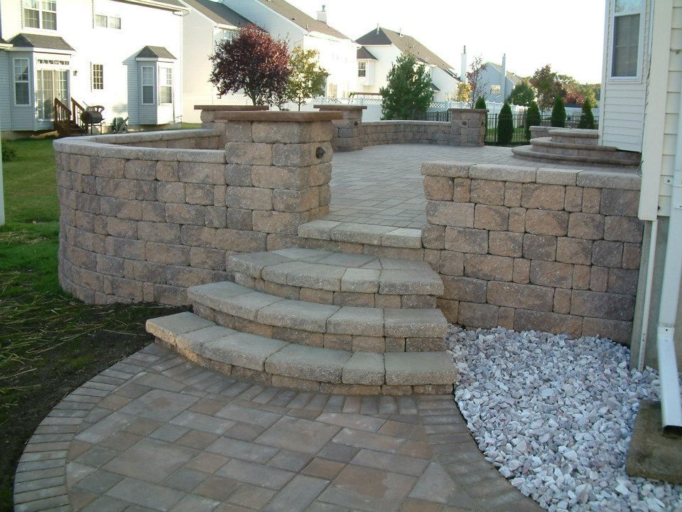 A backyard patio made of tan interlocking stone pavers with a curved retaining wall and semicircular tiered steps.
