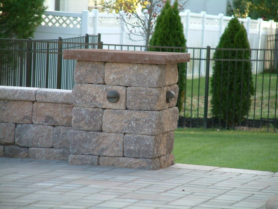 A stone pillar with two small lights sits on a patio next to a short wall, with a metal fence and shrubs in the background.