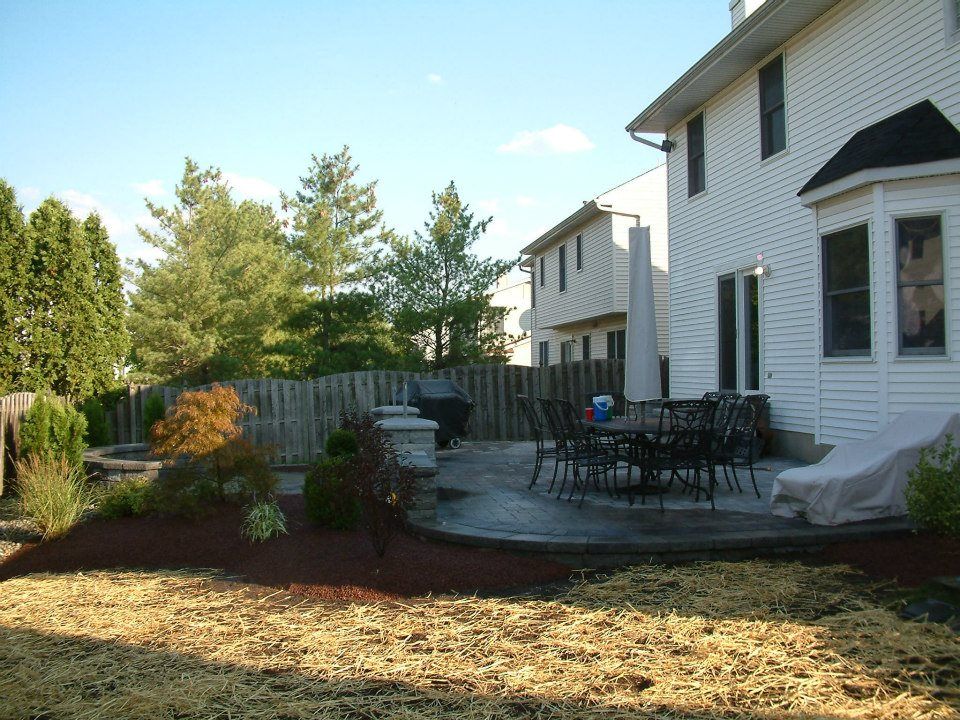 Backyard patio with a dining set and a covered lounge chair next to a white house with landscaping. Backyard patio with a dining set and a covered lounge chair next to a white house with landscaping.