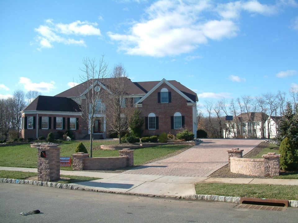 A large, two-story brick suburban home with a dark roof and paved driveway behind a stone-pillared gate. A large, two-story brick suburban home with a dark roof and paved driveway behind a stone-pillared gate.