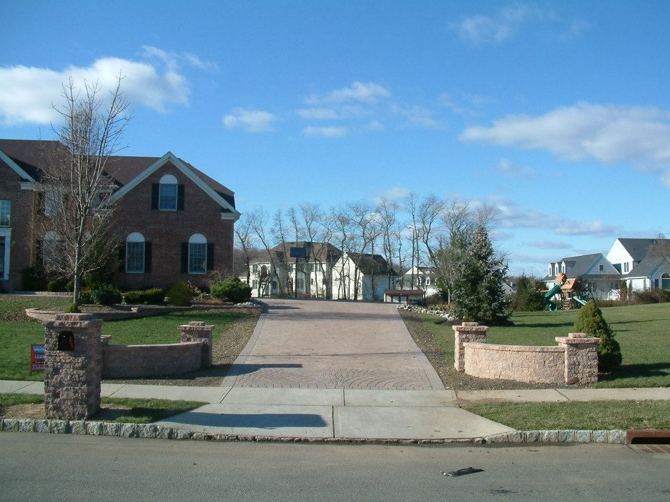 A long brick-paved driveway leads from a residential street toward a brick home under a clear blue sky.