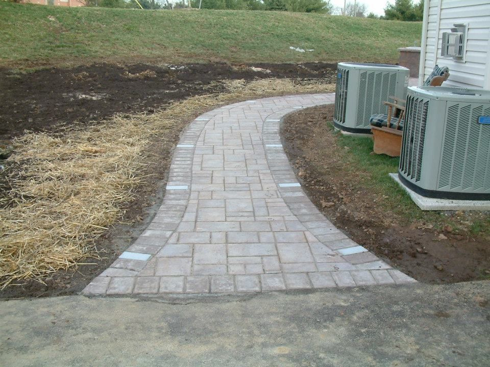 A stone paver walkway curves through a yard next to two grey outdoor air conditioning units and a house wall. A stone paver walkway curves through a yard next to two grey outdoor air conditioning units and a house wall.