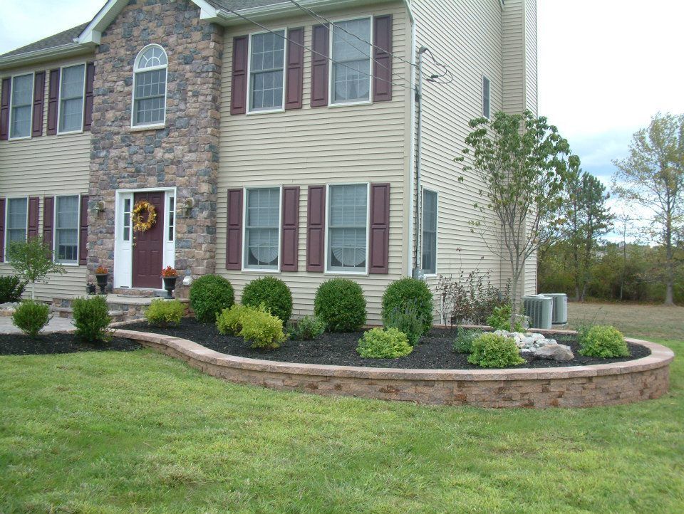A two-story tan house with a stone facade entrance features a curved stone retaining wall surrounding a landscaped bed.