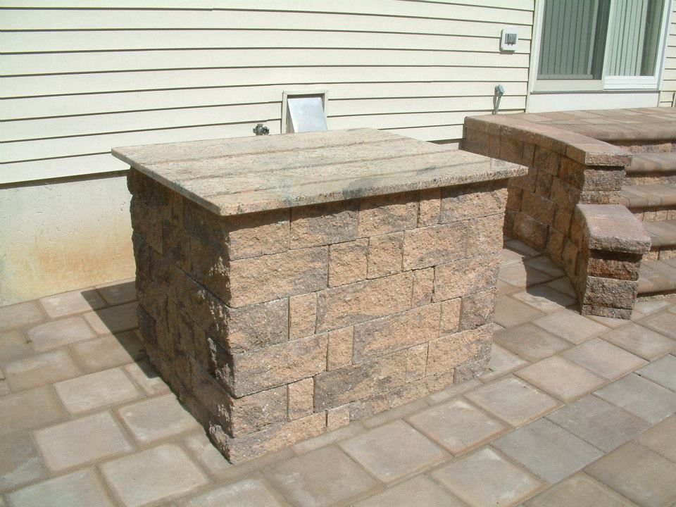 A stone outdoor island with a granite countertop sits on a paver patio next to a staircase and beige house siding.