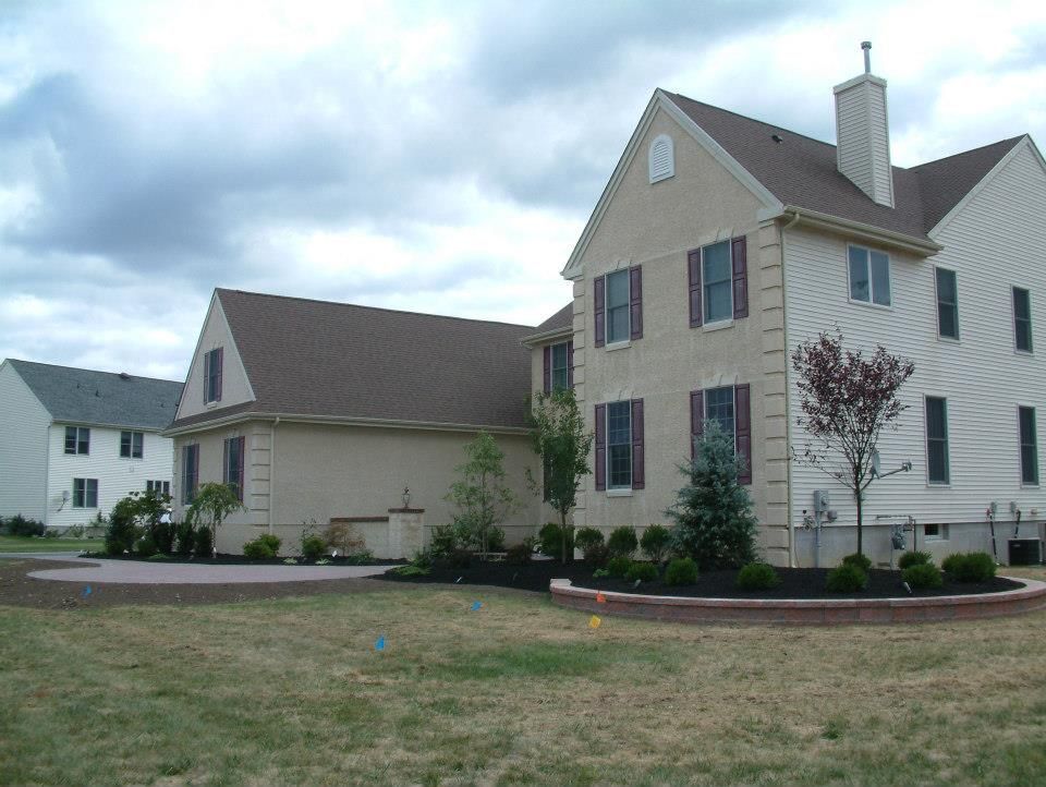 A two-story beige house with a brown roof and brown shutters, featuring a newly landscaped garden in the front yard.
