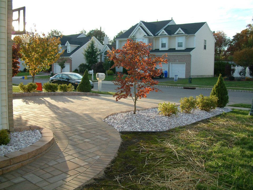 A paved walkway curves toward a front entrance, flanked by a landscaped garden bed with white stone, shrubs, and a tree.