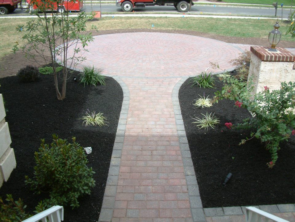 A brick pathway leads from a home's stairs to a circular patio, bordered by dark mulch and landscaping in a front yard.