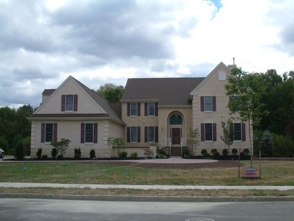 A two-story tan suburban house with dark shutters, a front entryway with an arched window, and a surrounding lawn.