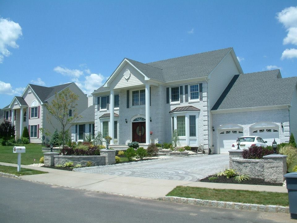 Two large, light-colored colonial-style houses with gray roofs, stone pillars, and manicured lawns under a clear blue sky.