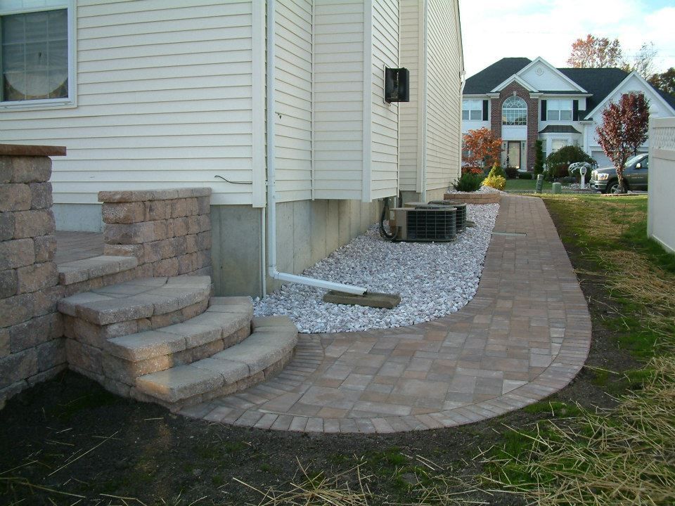Stone steps lead from a patio to a paved walkway alongside a white house with a gravel garden bed and AC unit.