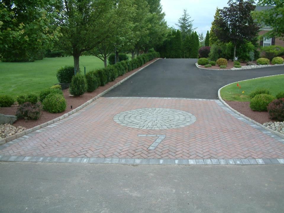 A paved driveway entrance featuring red herringbone brickwork with a circular gray stone pattern, bordered by shrubs.